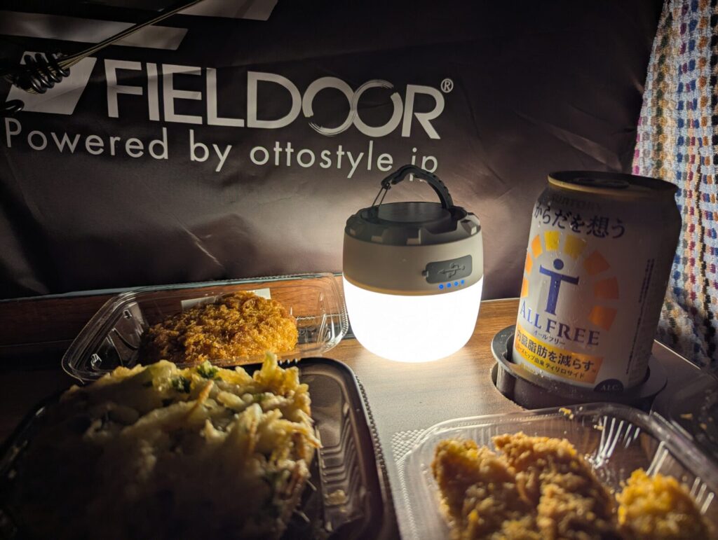 FIELDORのタープの下、LEDランタンの灯りに照らされた夜の車中飯。惣菜の揚げ物やかき揚げ、ノンアルコールビール『オールフリー』が並び、静かなアウトドアの時間を演出している。