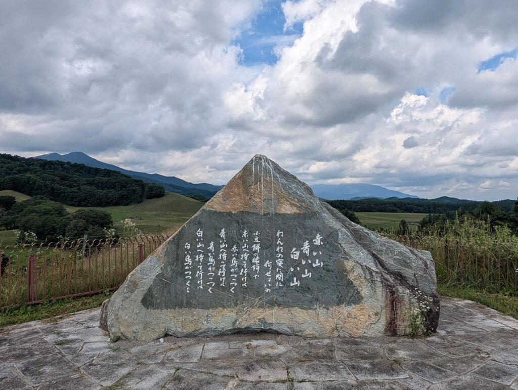 八千代牧場に建つ石碑。山並みと広大な牧草地を背景に、「赤い山 青い山 白い山」と刻まれた詩が大きな岩に刻まれている。